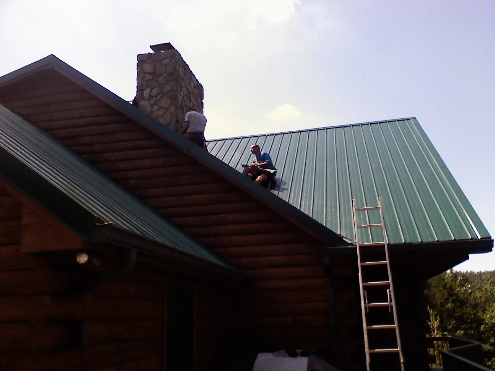 Roofers installing green metal panels beside a stone chimney on a log home in North Brunswick